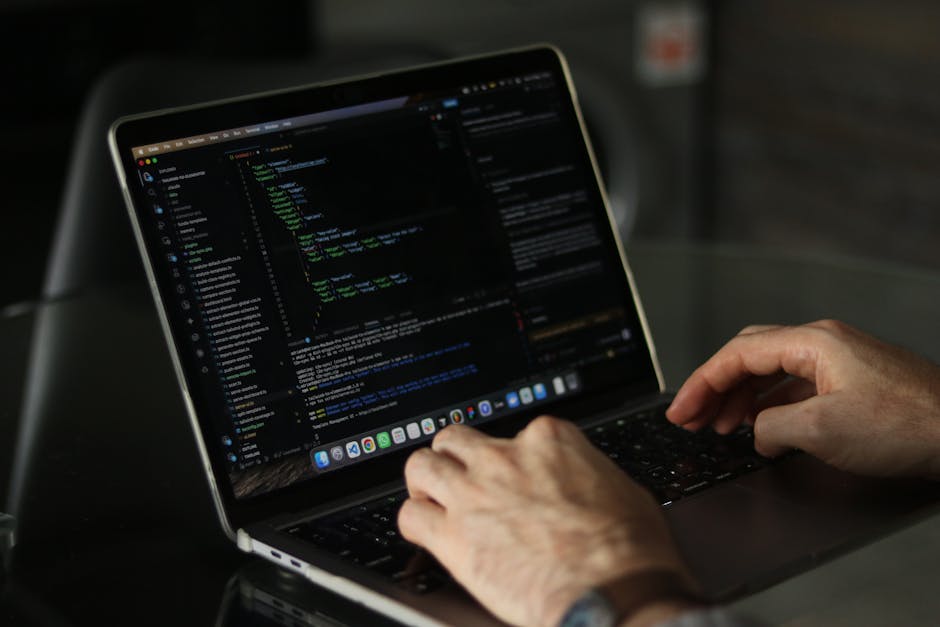 A close-up view of a person's hands operating a MacBook laptop placed on a dark surface, displaying lines of computer code with colorful syntax highlighting on the screen. The background is slightly b