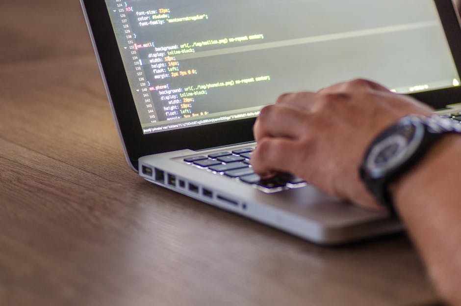 A close-up of a person's hand wearing a wristwatch, positioned over a silver laptop keyboard, with the screen displaying lines of colorful code on a dark background. The laptop is placed on a wooden s