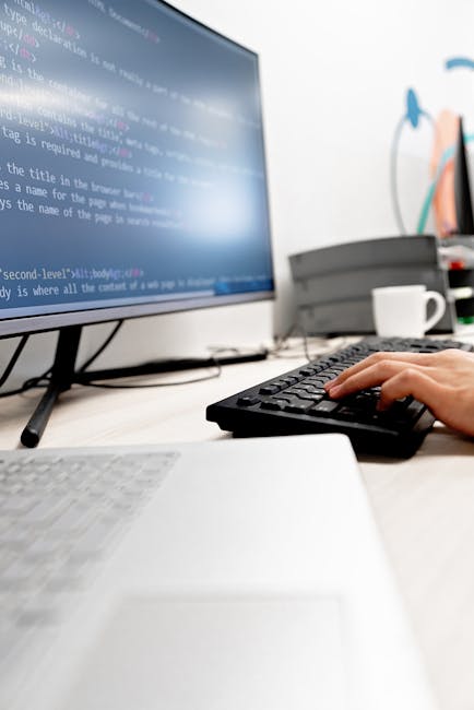 A close-up of a person's hand wearing a wristwatch, positioned over a silver laptop keyboard, with the screen displaying lines of colorful code on a dark background. The laptop is placed on a wooden s
